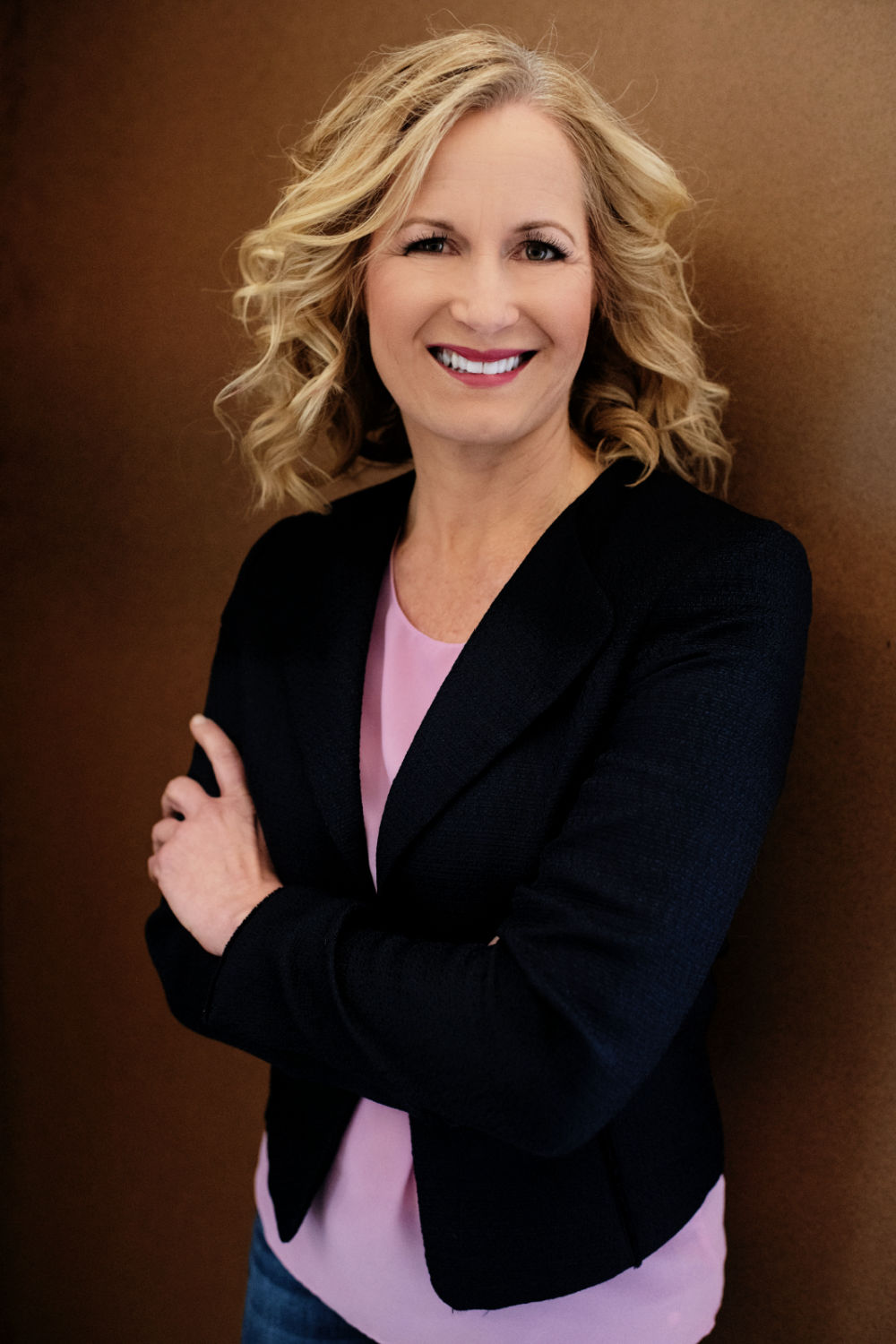 Jennifer smiling with her arms casually crossed, wearing a jacket and blouse over jeans